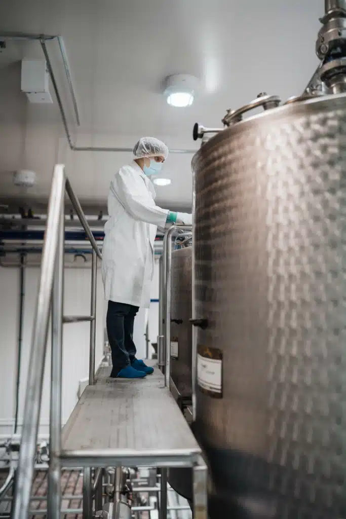 A worker in a modern dairy processing facility, wearing a white lab coat, hairnet, and mask, standing on a platform beside large stainless steel tanks.