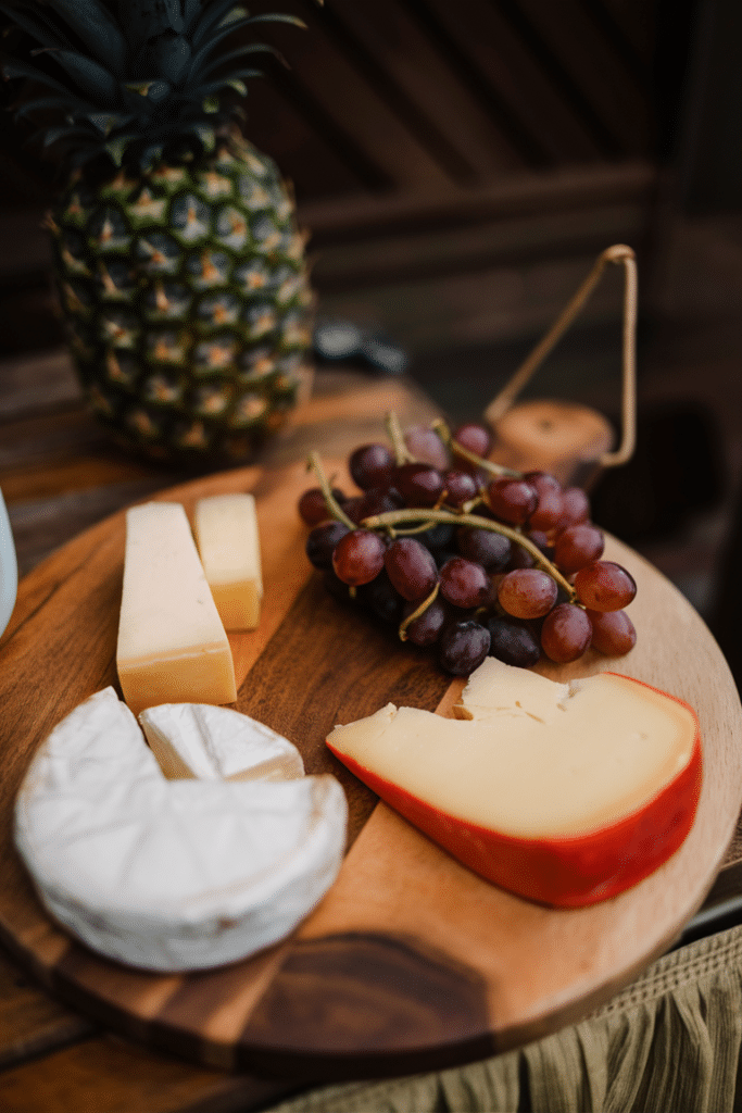 A cheese board featuring several cheese slices, a bunch of grapes, and a pineapple in the background.