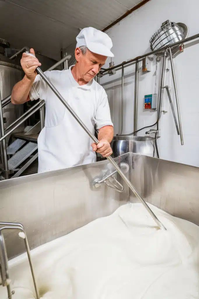 A cheesemaker stirring a large vat of milk in a modern cheese production facility, wearing a white uniform and cap, with stainless steel equipment in the background.