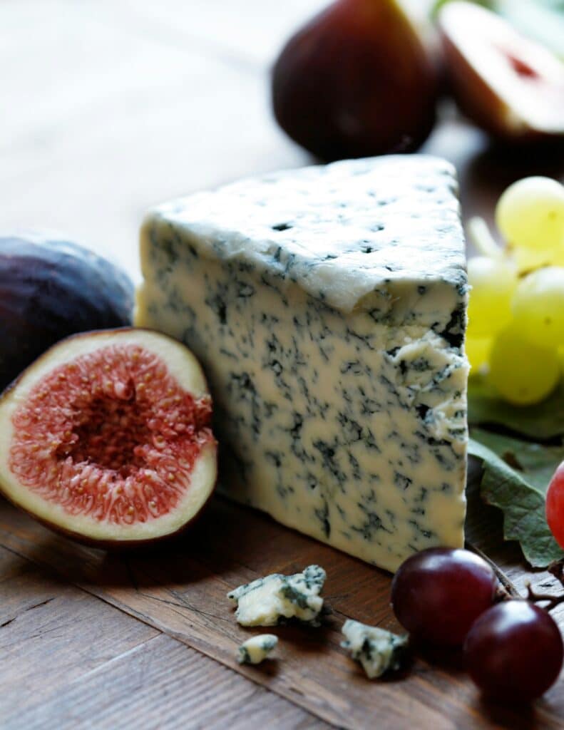 A wedge of blue cheese with a creamy texture and blue mold veins, accompanied by fresh figs, grapes, and a peach on a wooden surface. The scene suggests a rustic, gourmet cheese platter setting.