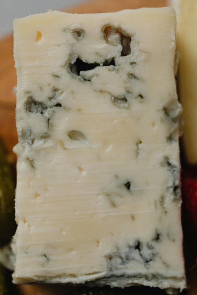 A close-up view of a block of blue cheese, highlighting its creamy texture and blue-green mold veins. The cheese is set against a blurred background, with a hint of other food items visible.