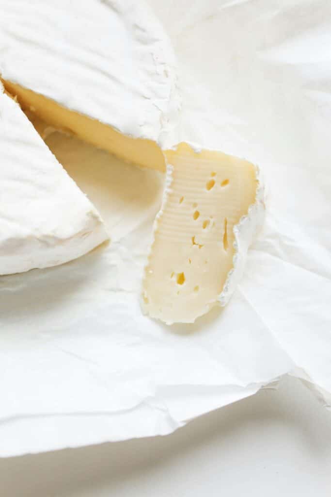 A close-up view of a wedge of Brie cheese cut from a whole wheel, resting on white parchment paper. The cheese displays a creamy interior with a few small holes and a smooth, white rind, emphasizing its soft and velvety texture.