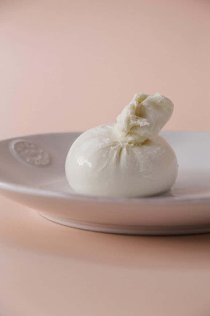 A close-up view of a ball of Burrata cheese, neatly tied at the top, sitting on a white plate. The background is a soft, peach-colored surface, highlighting the cheese's smooth, glossy texture and fresh appearance.