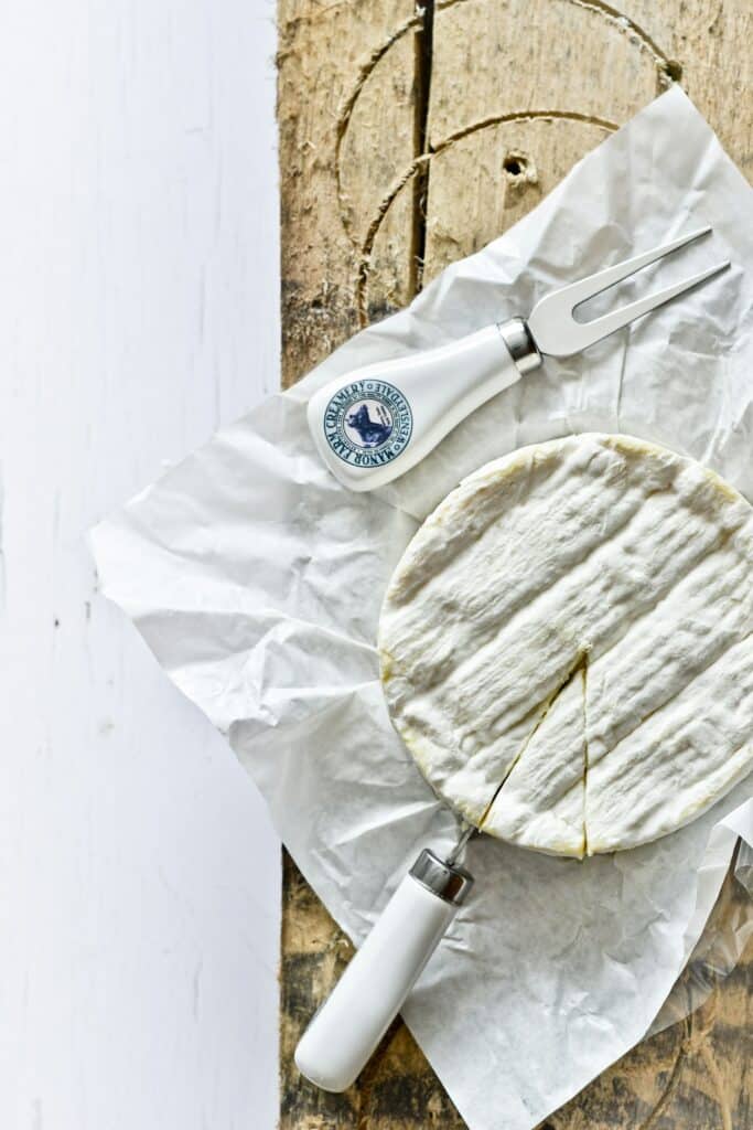 A top-down view of a whole wheel of Camembert cheese with a small wedge cut out, resting on white parchment paper. A cheese knife with a white handle is placed beside the cheese on a rustic wooden board, highlighting the cheese's textured rind and creamy interior.