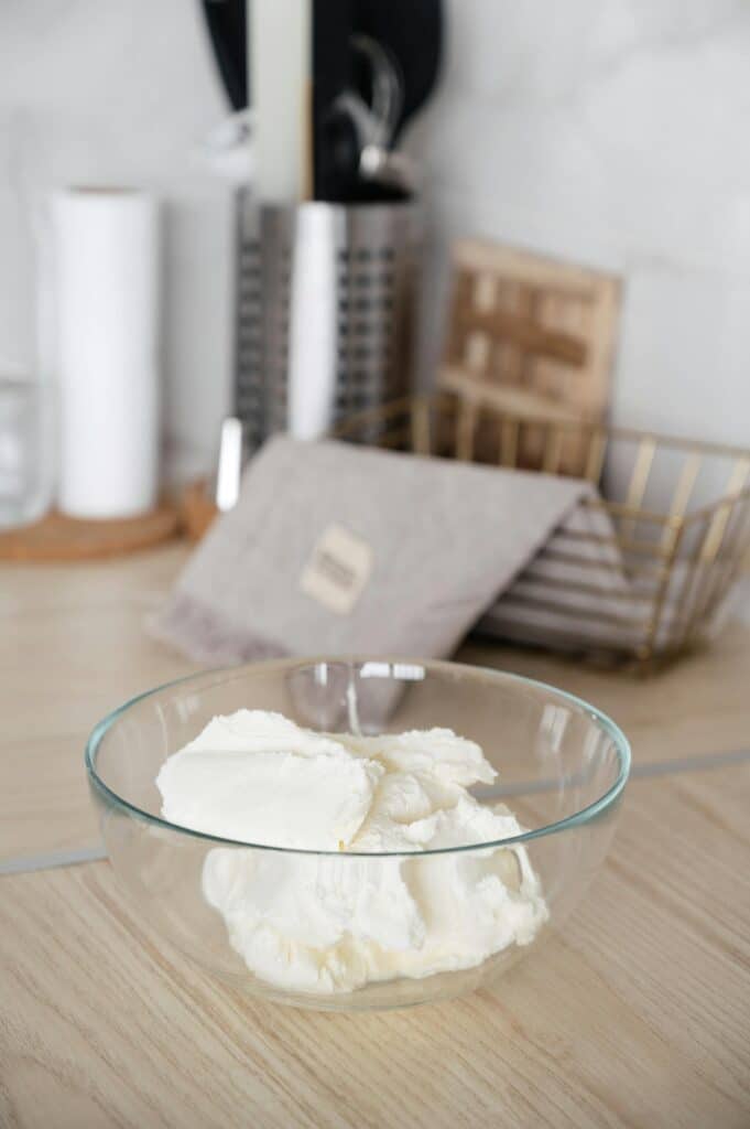 A glass bowl filled with cream cheese, placed on a wooden kitchen countertop with various kitchen utensils and a dish rack in the background.