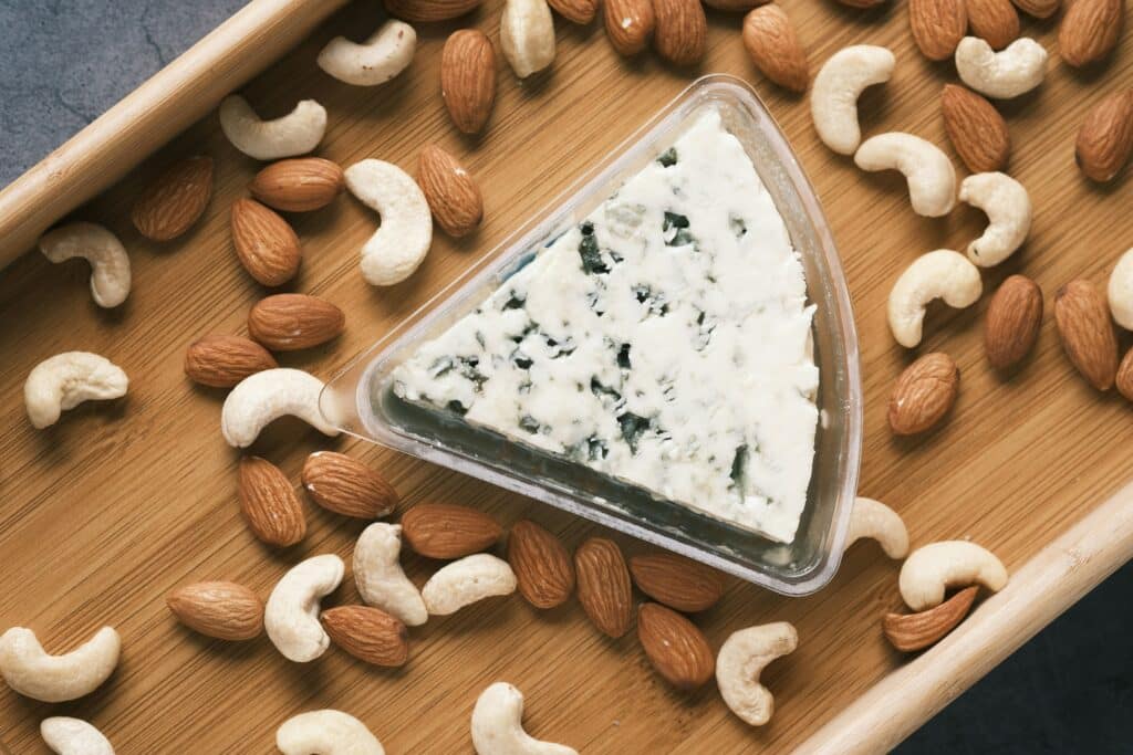  A wedge of blue cheese in a plastic container, placed on a wooden tray surrounded by scattered almonds and cashews. The scene suggests a snack or appetizer setup, highlighting the combination of cheese and nuts.
