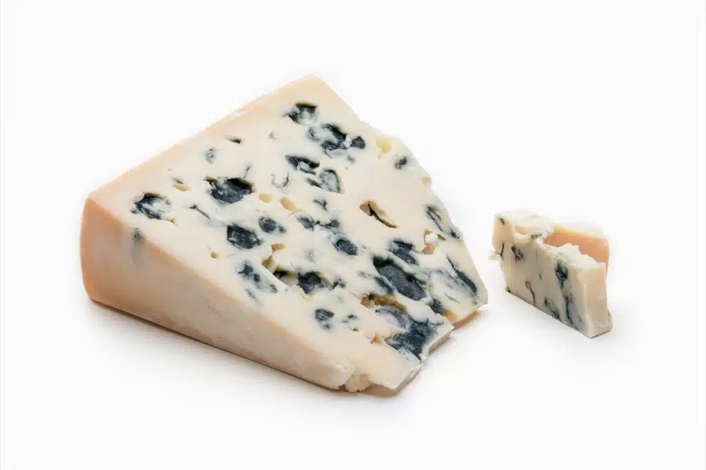 A wedge of Roquefort cheese with a creamy white texture and distinctive blue mold veins, accompanied by a small chunk broken off from the main piece. The cheese is set against a white background.