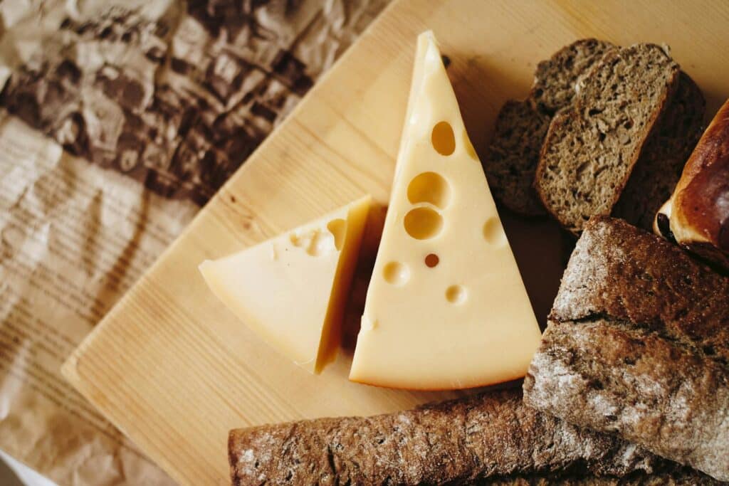 A wedge of Swiss cheese with characteristic holes, accompanied by slices of rustic bread, arranged on a wooden cutting board.