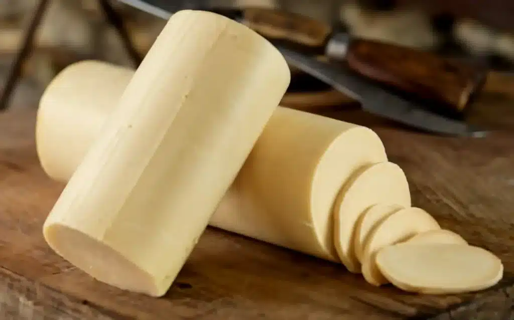 A photo of a wooden table with a provolone cheese. Adjacent to the cheese, there are slices of the same object, neatly arranged. In the background, a vintage knife with a wooden handle can be seen, suggesting that the object was recently sliced. The overall ambiance of the image is rustic, emphasizing the natural and organic nature of the product. The background is blurred, showing a rustic setting with stone walls and wooden elements.