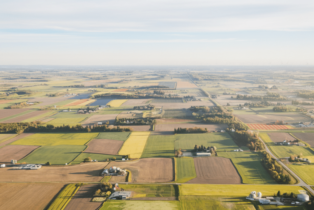 Aerial view of vast farmland in Canada, with a patchwork of green, brown, and yellow fields, scattered farm buildings, and distant roads stretching towards the horizon under a clear sky.