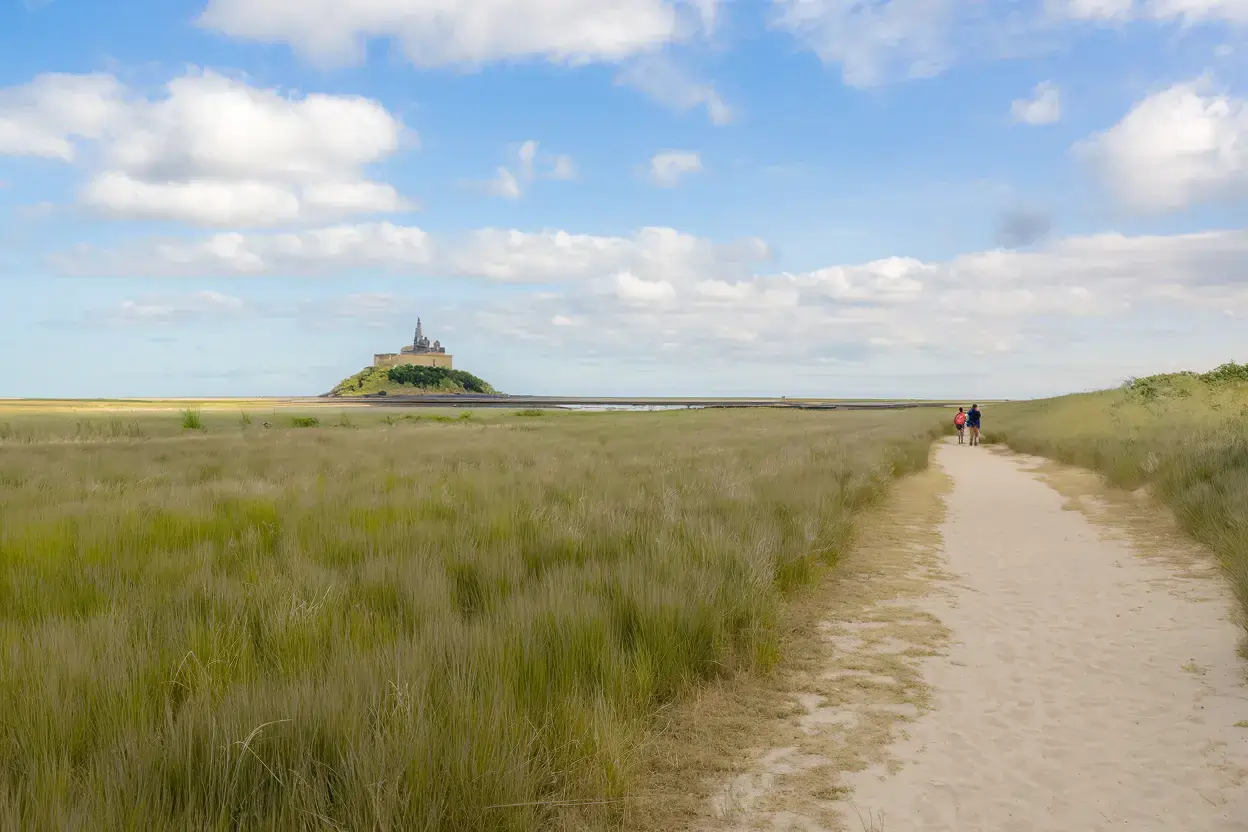 A vast, open landscape with a sandy pathway leading towards a distant structure. On the left, there's a field of tall grasses, and on the right, there's a grassy hill. The sky is blue with scattered white clouds. In the distance, there's a prominent structure, possibly a historic building or monument, situated on a small island or elevated landmass. A few people can be seen walking on the pathway, giving a sense of scale to the vastness of the surroundings.